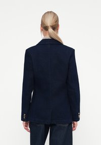 Marineblå blazer med skreddersydd passform, skjærte revers og tre gullknappeakenter på hver mansjett, laget av et strukturert stoff.