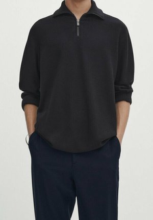 Homme portant un pull noir à fermeture éclair quart de zip avec les manches retroussées et un pantalon bleu marine, les mains dans les poches, debout devant un fond uni.