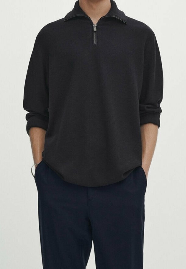 Homme portant un pull noir à fermeture éclair quart de zip avec les manches retroussées et un pantalon bleu marine, les mains dans les poches, debout devant un fond uni.