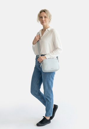 Sieviete baltā kreklā un zilās džinsās stāv, turot gaiši pelēku plecu somu, valkā melnas sporta apavus uz baltas fona.