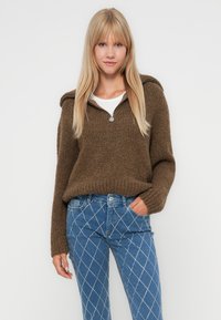Brun strikket genser med glidelås i kragen, kombinert med blå jeans med diamantmønster og pynt, vist på en nøytral bakgrunn.