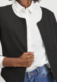 Sort tekstureret jakke med lynlås foran, båret over en hvid skjorte med knapper og dekorativ søm i kanten. Blå denim ses nedenfor.