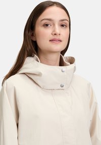 Młoda kobieta o prostych brązowych włosach, ubrana w beżową kurtkę z wysokim kołnierzem zapinaną na zatrzaski, na tle jednolitego jasnego tła.
