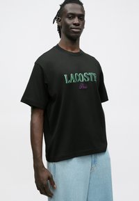 T-shirt de algodão preta com corte solto, apresentando o texto bordado em verde e roxo "LACOSTE Paris" no peito. Gola redonda padrão.