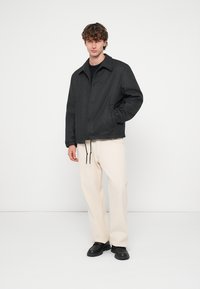 Šedá, textúrovaná bunda s golierom, prednými vreckami a sťahovacou šnúrkou. Kombinovaná s béžovými nohavicami a čiernymi topánkami. Jednofarebné pozadie.