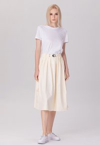 T-shirt blanc associé à une jupe longueur genou de couleur jaune pâle, avec des plis et une ceinture, le tout complété par des baskets blanches.