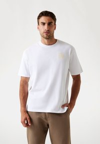 Hvid bomuld t-shirt med rund hals og korte ærmer. Har et kvadratisk logo-patch på venstre bryst. Blød tekstur, afslappet pasform.