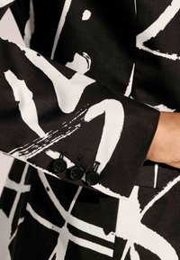 Zwarte stof met gedurfde witte abstracte patronen, met een op maat gemaakte manchette met drie zwarte knopen en verhoogde stikdetail.