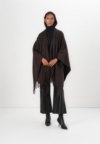 Brauner Poncho aus weichem Stoff, mit drapiertem Design und fransigen Kanten. Getragen über einem schwarzen Rollkragenpullover und kombiniert mit schwarzen Hosen.