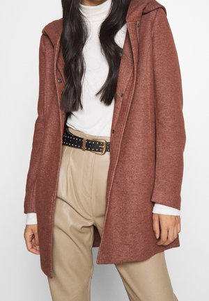 Femme portant un manteau couleur rouille par-dessus un col roulé blanc, un pantalon beige taille haute et une ceinture noire avec des clous dorés et une boucle.