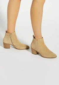 Beige suede ankle boots with a pointed toe, side zipper, and a stacked heel. Smooth texture with minimal stitching details.