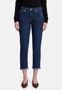 Jeans in denim blu scuro con una vestibilità slim e orli alla caviglia, dotati di cinque tasche e chiusura con cerniera, indossati con scarpe marroni a fantasia.