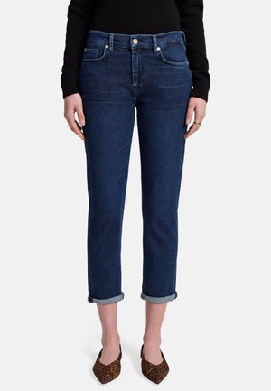 Jeans in denim blu scuro con una vestibilità slim e orli alla caviglia, dotati di cinque tasche e chiusura con cerniera, indossati con scarpe marroni a fantasia.