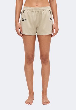 Femme portant un short UFC beige avec une taille élastique et un logo noir à l'avant, debout pieds nus devant un fond uni.