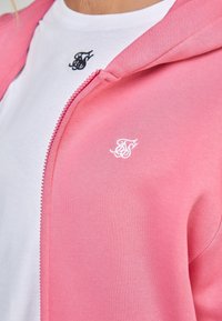 Roze ritshoodie van zacht materiaal, met een subtiele witte logo op de borst, gecombineerd met een witte t-shirt die eronder zichtbaar is.