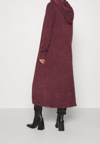 Cardigan con cappuccio lungo color marsala, texture morbida, vestibilità ampia, arriva fino al polpaccio. Abbinato a stivaletti neri con tacco e jeans scuri.