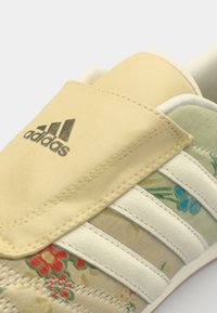 Boty Adidas s květinovým textilním svrškem v béžové a zelené barvě, s bílými koženými pruhy a žlutým textilním jazykem, logo na jazyku.