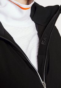 Black jacket with a zip closure, featuring a white textured inner layer, and a high collar with an orange accent stripe.