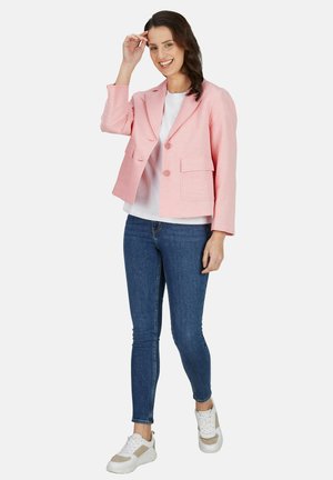 Glimlachende vrouw die een roze blazer, wit overhemd, blauwe skinny jeans en witte sneakers draagt, staand en haar haar aanrakend.