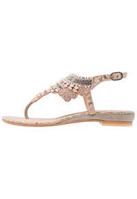 Beżowe płaskie sandały z ozdobnymi koralikami i elementami z kryształów, z paskiem wokół kostki z klamrą oraz subtelną teksturowaną podeszwą.