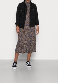 Giacca di denim nera sopra un vestito midi floreale con motivi blu e beige, abbinato a sneakers bianche e nere, in piedi su un pavimento grigio chiaro.