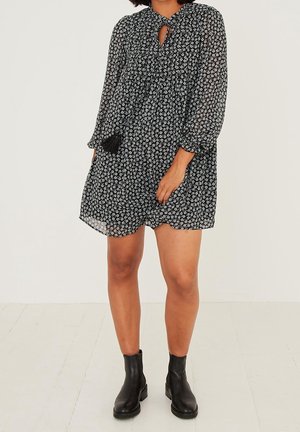 Femme portant une robe longue à manches longues noire et blanche à motif floral, associée à des bottines noires, debout sur un sol clair devant un arrière-plan uni.