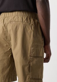 Pantaloni cargo color khaki con vita elasticizzata, tessuto testurizzato, tasche laterali e tasca applicata sulla coscia.