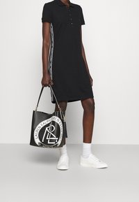 Svart poloklänning med korta ärmar och guldknappar, med vit sidobrev. Komplementeras av en svart tote-väska med en cirkulär logotyp.