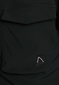 Prenda de tela negra con un bolsillo de solapa prominente, que presenta una etiqueta tejida rectangular con un logo blanco. Textura suave y diseño minimalista.