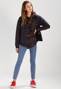 Svart, polstret jakke med hette, trykknapper, brystlomme; brukt over en mørk camo print hettegenser, paret med blå jeans og røde sneakers.