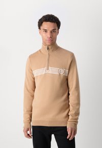 Beżowy sweter na zamek z półkołnierzykiem, z białym paskiem na piersi i okrągłym logo po lewej stronie.