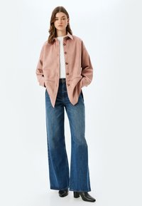 Rosa, mockasinsliknande knäppt jacka med krage, två framfickor och sidoflikar, kombinerad med vida blå denimjeans och svarta ankstövlar.