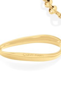 Pulsera de metal tono dorado con un diseño liso y redondeado. Presenta el logotipo grabado de "Calvin Klein" en la superficie.