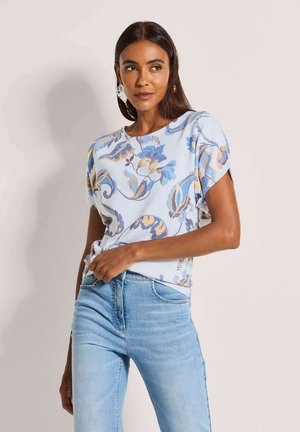 Frau trägt ein hellblaues, kurzärmliges Oberteil mit Blumenmuster und hellblaue Jeans, posiert mit der Hand in der Hosentasche.