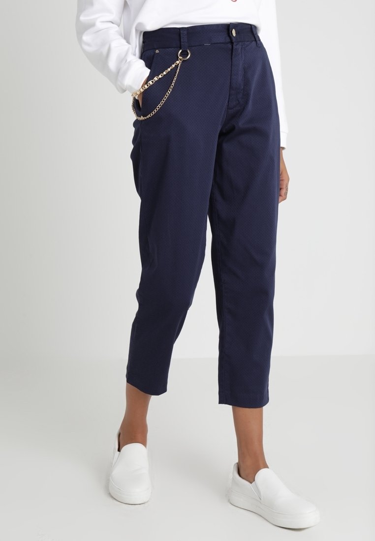 Pantalon bleu marine fuselé avec longueur raccourcie, accessoire chaîne dorée sur la poche, associé à des chaussures blanches slip-on et un haut blanc.