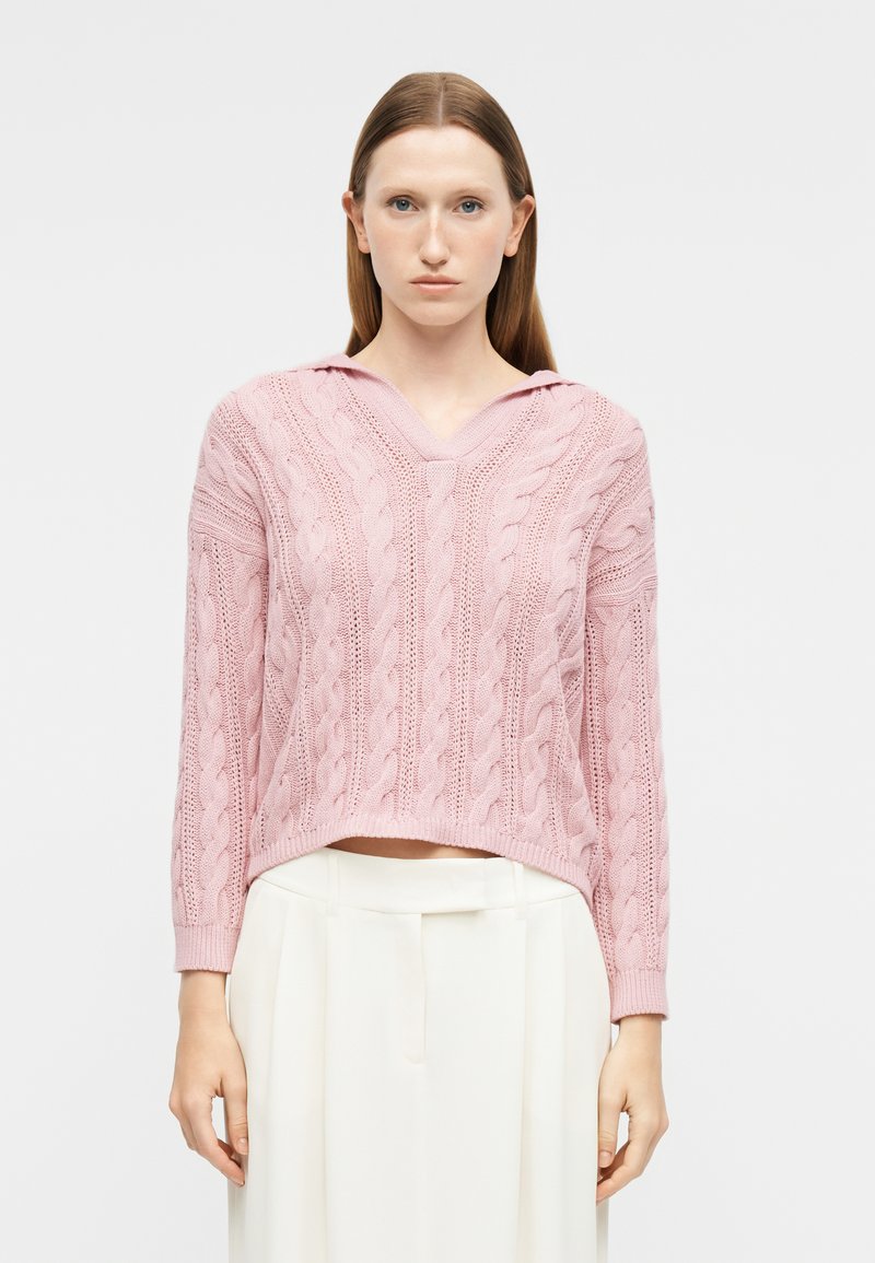 Femme portant un pull en maille torsadée rose clair avec un col en V et un pantalon blanc taille haute, sur un fond uni.