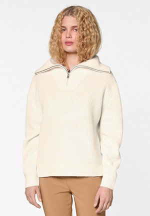 Femme aux cheveux blonds bouclés portant un pull crème côtelé avec un col zippé et un pantalon marron clair sur un fond uni.