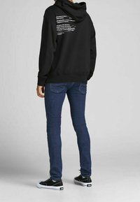 Person standing, wearing dark blue skinny jeans, black hoodie with white text on back, and black low-top sneakers with white soles.