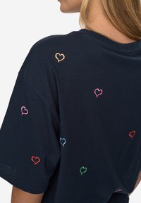 Camiseta azul marino con bordados de corazones multicolores en las mangas y la espalda. Mangas cortas, ajuste holgado, textura suave.