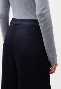 Calças largas azul-marinho com textura de nervuras diagonais e detalhe de bolso traseiro, combinadas com uma blusa justa de manga longa cinza.