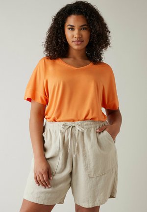 Frau mit lockigem Haar, die ein oranges kurzärmeliges Oberteil und beige Shorts mit Kordelzug trägt, steht mit einer Hand in der Tasche.
