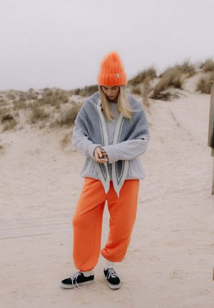 Młoda kobieta w jaskrawo pomarańczowej czapce i spodniach, szarym swetrze i szalu, stoi na piaszczystych wydmach, patrząc w dół na swoje dłonie.