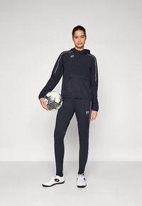 Tumesinine sportlik komplekt, mis koosneb kapuutsiga pulloverist valgete detailidega ja sobivatest, hästiistuvatest pükstest. Modell hoiab must-valget jalgpalli.