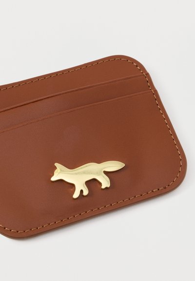 Καφέ δερμάτινη θήκη καρτών με χρυσό έμβλημα αλεπούς. Διαθέτει δύο υποδοχές για κάρτες και ορατές ραφές γύρω από τις άκρες. Απαλή υφή.