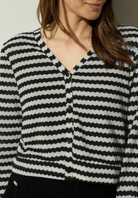 Černobílý pruhovaný svetr s výstřihem do V, který má strukturální cikcak vzor a černé knoflíky na přední straně. Měkký pletený materiál.