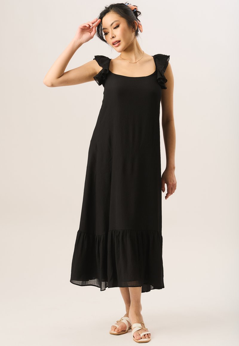 Black sleeveless dress with ruffled shoulder accents, a relaxed fit, and a tiered ruffle hem, made from lightweight fabric. Flat sandals.