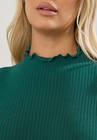 Top verde a costine con collettino alto e bordo arricciato. Il tessuto appare liscio e lucido con una silhouette aderente.