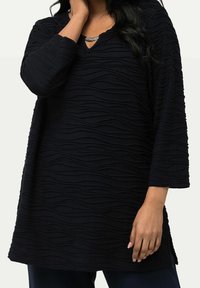 Svart tunika med texturerat vågmönster, V-ringning, trekvartsärmar och slitsar på sidorna för ökad rörelsefrihet.