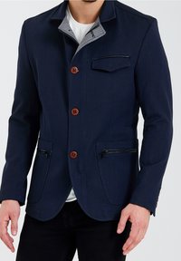 Marinblå jacka med strukturfinish, som har en grå krage, träknappar och två framfickor med dragkedja. Skräddarsydd passform.