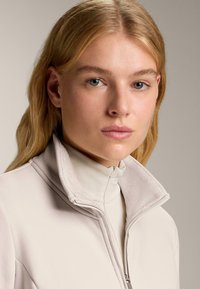 Tânără cu păr blond și ochi albaștri poartă o jachetă bej deschis, cu fermoar și guler înalt, pe un fundal simplu.